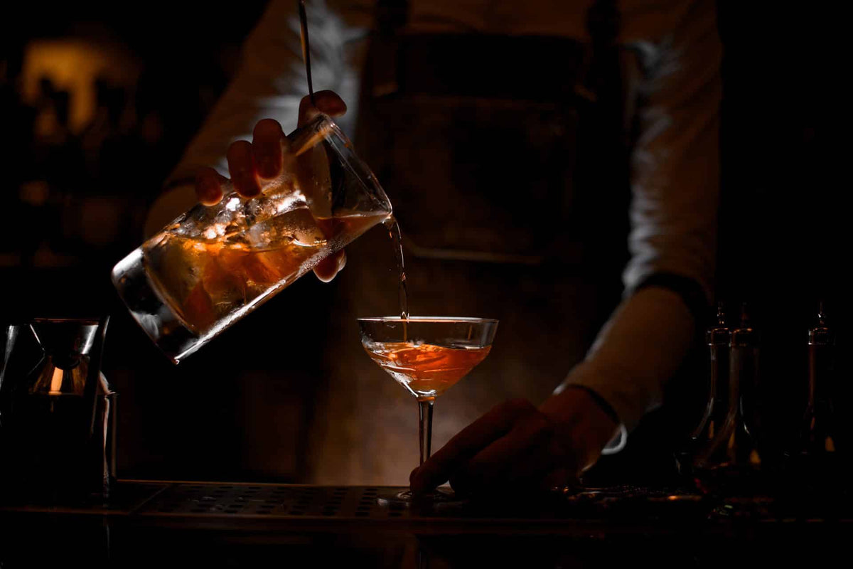 A bartender in a dim bar setting pours an amber-orange cocktail from a glass measuring pitcher into a coupe glass.