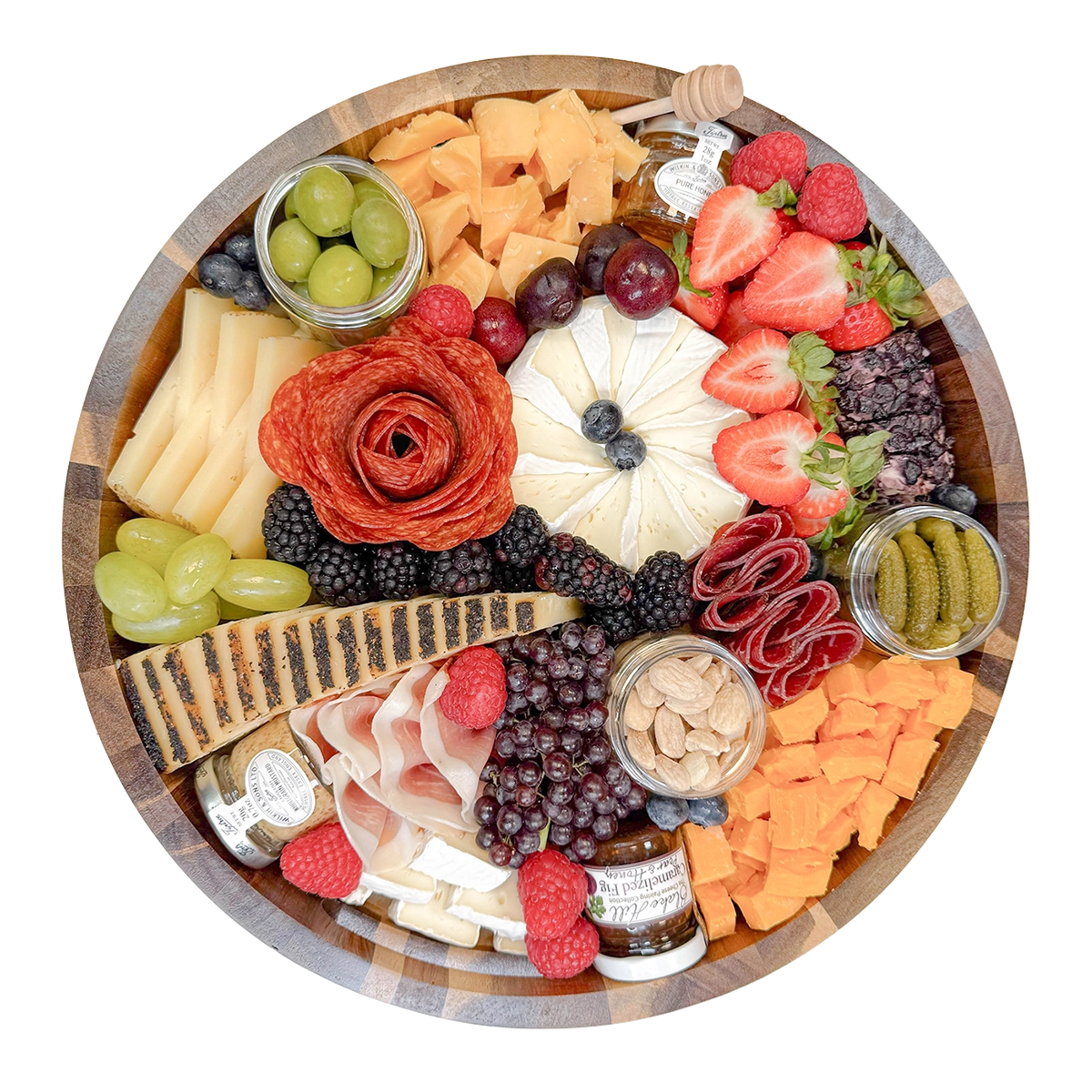 A round wooden charcuterie board arranged with cheeses, crackers, sliced meats, fruit, pickles, nuts, and small jars on a white background.