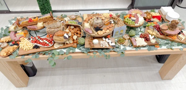 A multi-dimensional corporate grazing table by Fork and Flare in Irvine, California, featuring an abundant spread of salami, prosciutto, gouda, brie, dill, port wine cheddar, merlot, manchego, and boursin cheeses, paired with caprese skewers, artisan bread, crackers, pistachios, cornichons, grapes, oranges, white and orange cheddar, and finished with brownie bites and colorful macarons for a luxurious presentation.